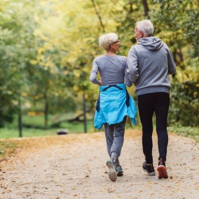 two people with grey hair are walking away from the camera along a sandy track with green vegetation either side